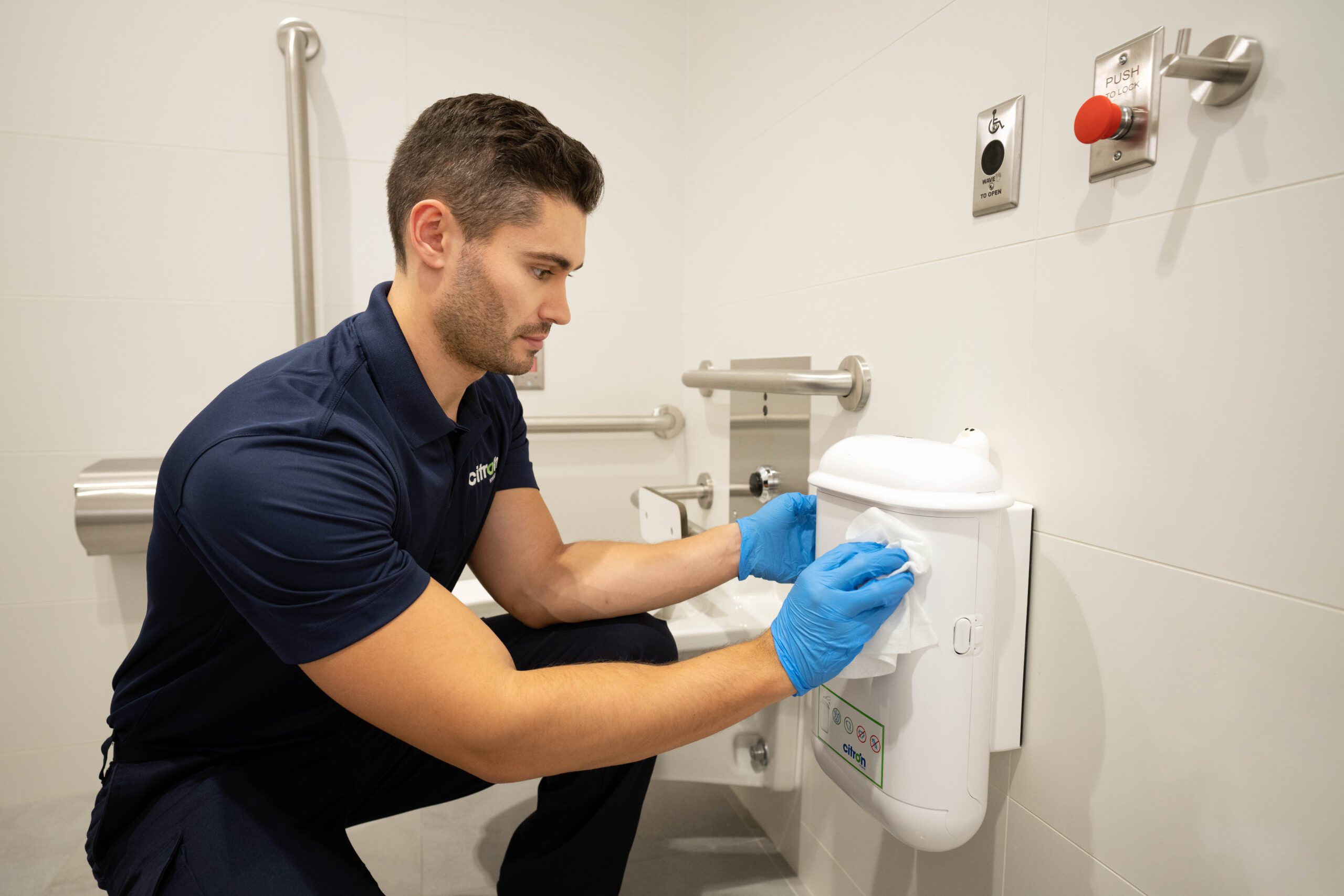 Cleaning a wall-mounted hygiene dispenser in a public washroom—representing Citron Hygiene’s commitment to safe, sanitary menstrual waste disposal.
